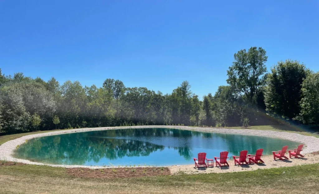 Better Way Land Management completed natural swimming pond with red Adirondack chairs on sandy beach area surrounded by mature trees
