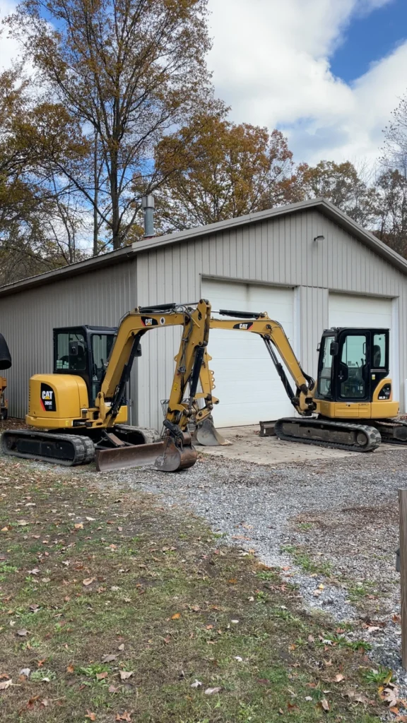 Better Way Land Management LLC CAT mini excavators with buckets parked on gravel near metal building in autumn equipment yard.